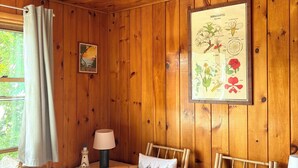 Une pièce chaleureuse aux murs et au plafond en bois, deux fauteuils en osier avec des coussins, une petite table avec une lampe et, au mur, une illustration botanique encadrée.