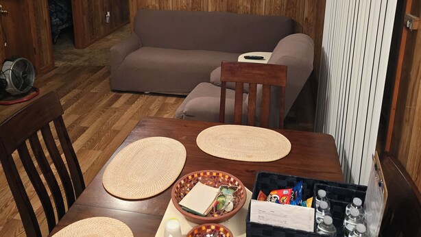 Un salon chaleureux avec un plancher en bois, un canapé marron, une table à manger avec des sets de table, un ventilateur de plafond et un tableau représentant un paysage, encadré.