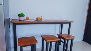 A small kitchen area with a wooden table, two stools, a potted plant, and a flat-screen TV mounted on the wall.