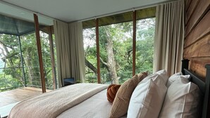 A bedroom with a large bed, multiple pillows, a view of trees through large windows, and a balcony.