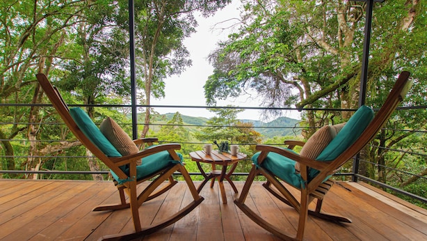 A wooden deck with two rocking chairs and a small table, overlooking a lush landscape with trees and hills.