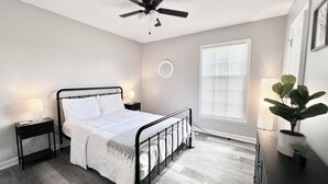 Een slaapkamer met een bed, een plafondventilator, een nachtkastje met een lamp, een raam met jaloezieën en een kamerplant.