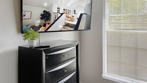 Een moderne kamer met een grote flatscreen televisie aan de muur, een zwarte dressoir met lades, een potplant en een raam met zonwering.