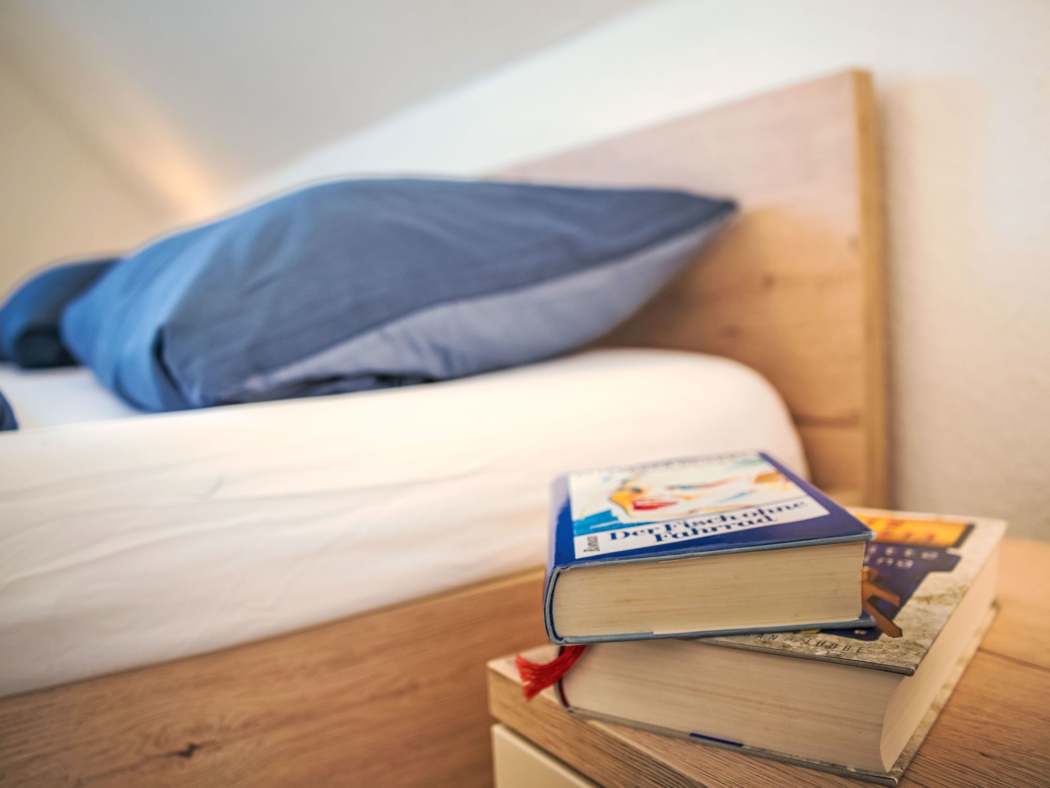 Un llit ben fet amb un coixí blau i una pila de llibres sobre una tauleta de nit de fusta.