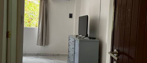 A room with a marble floor, a grey cabinet with a flat-screen TV, a window with curtains, and an air conditioning unit on the wall.