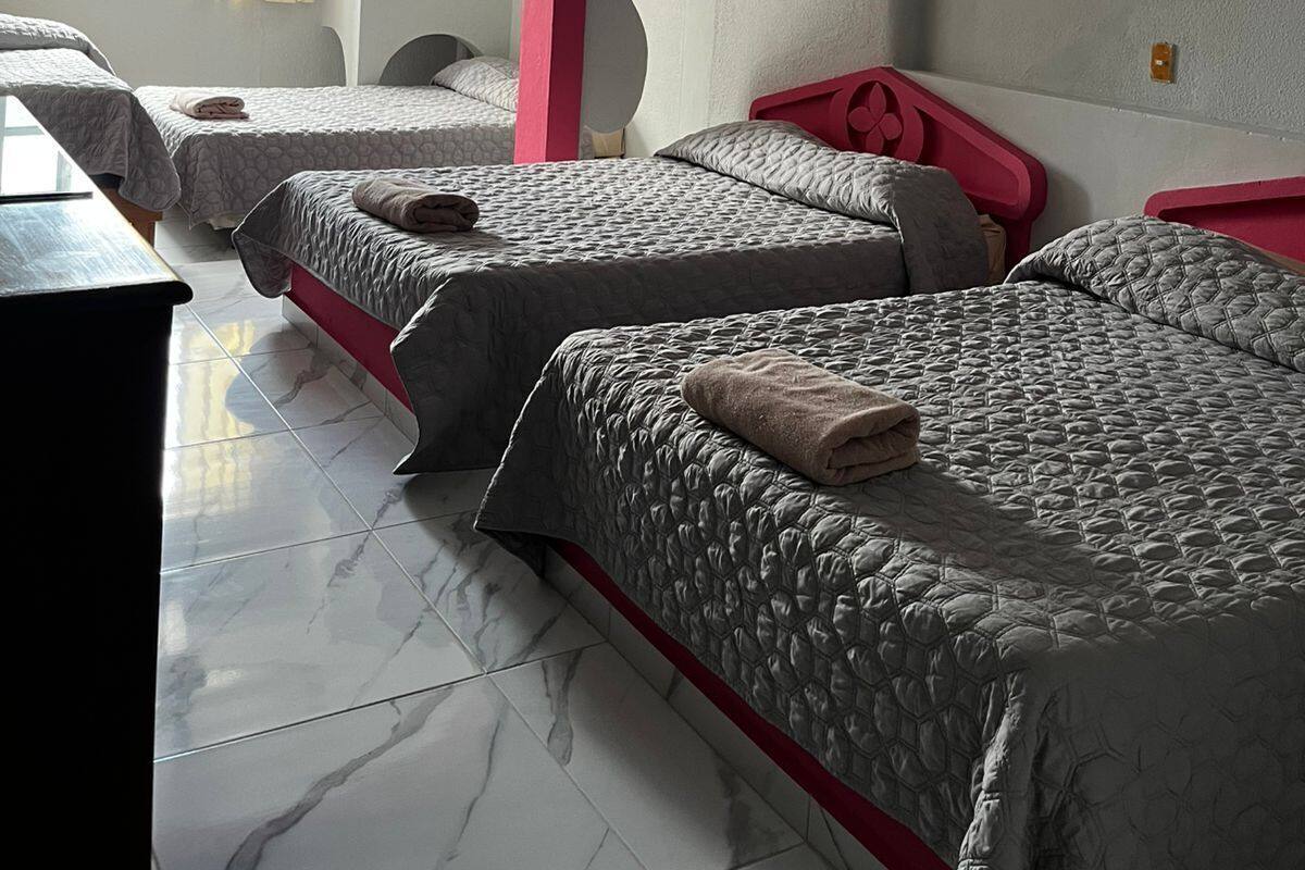 Habitación de hotel con tres camas, ventilador de techo, ventana con cortinas y lámpara fijada a la pared.