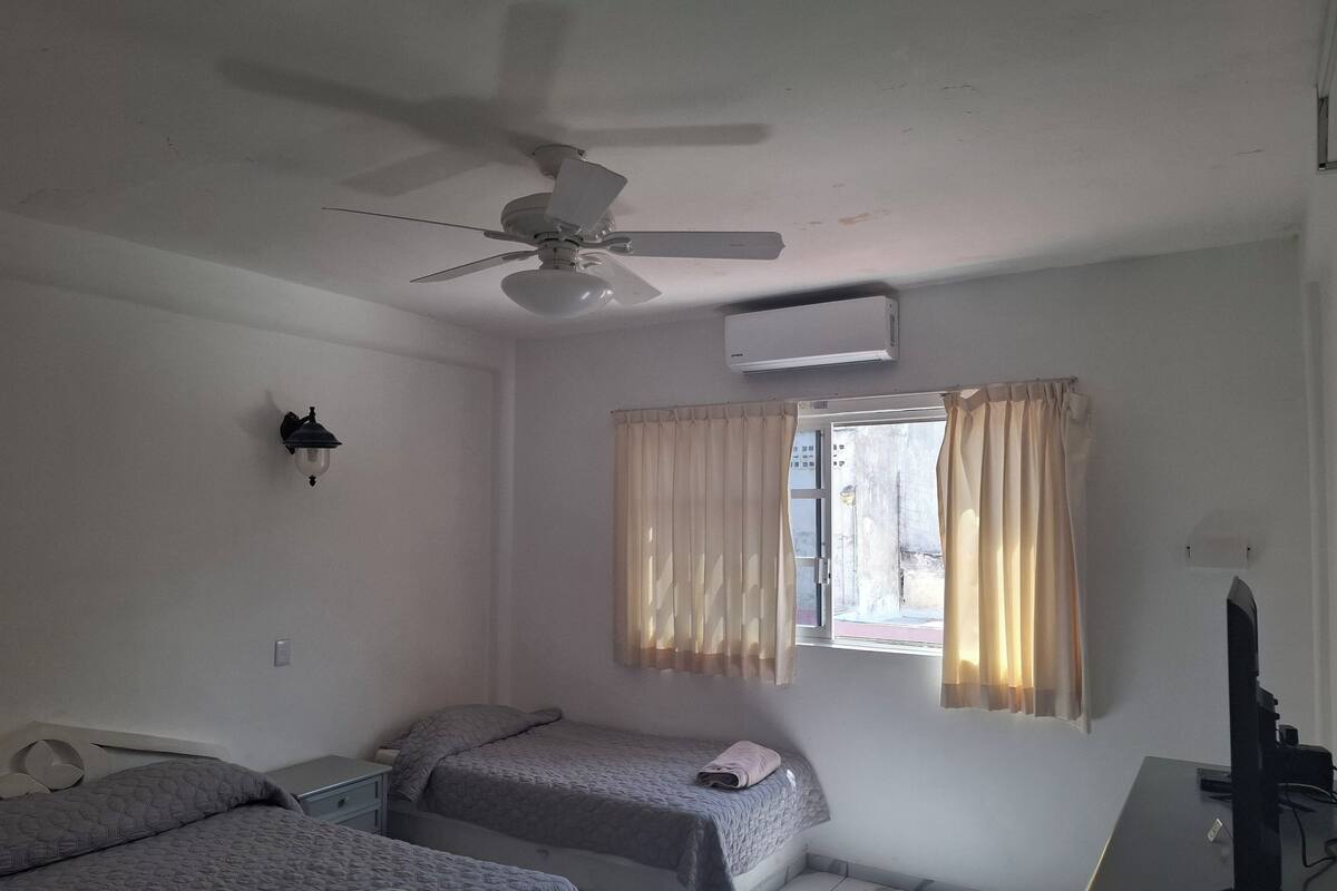 Una habitación con dos camas, ventilador de techo, aire acondicionado, escritorio con ordenador y ventana con cortinas.