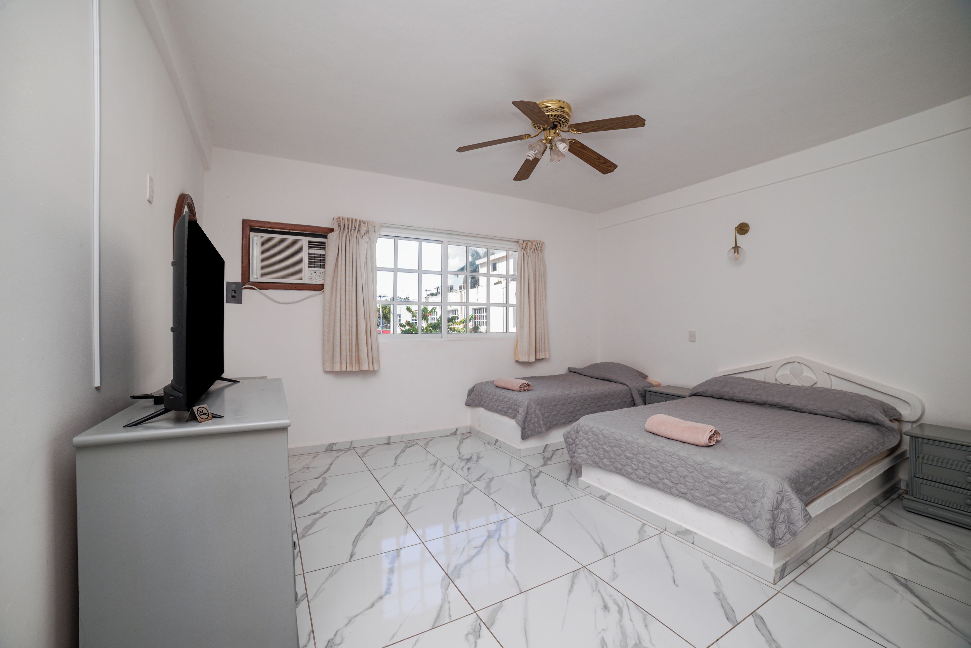 A bedroom with two beds, a ceiling fan, a window with curtains, and a television on a cabinet.