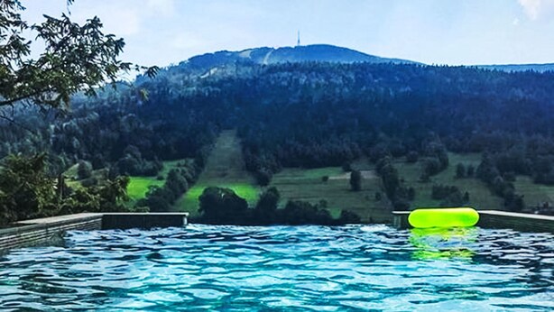 Une piscine à l’eau d’un bleu cristallin, entourée d’une terrasse en bois et offrant une vue sur une colline boisée.