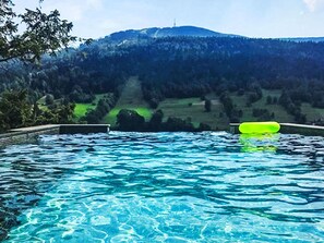 Una piscina con agua azul cristalina, rodeada por una terraza de madera y vista a una ladera boscosa.