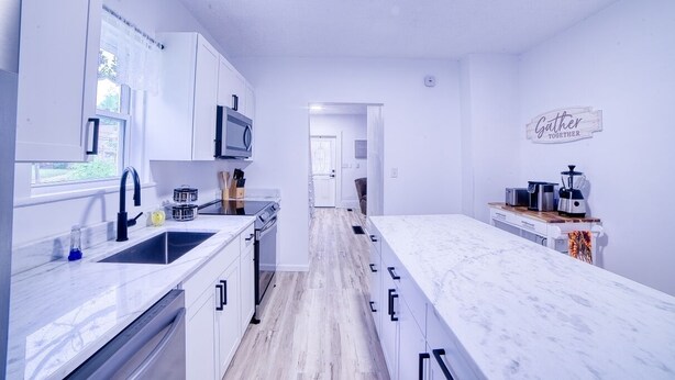 Una cocina moderna con armarios blancos, encimera de mármol y una isla central. Hay un microondas sobre el fregadero, una cafetera en la encimera y un letrero en la pared que dice “Gather Together”.