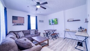 Una sala de estar con sofá, mesa de centro y televisor. Hay un ventilador de techo, una ventana con cortinas y un televisor fijado a la pared.