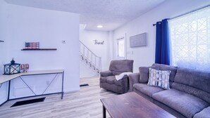 Una sala de estar moderna con un sofá, un sillón y una mesa de centro. Un escritorio con lámpara y objetos decorativos está contra la pared. Una escalera conduce a otro nivel.