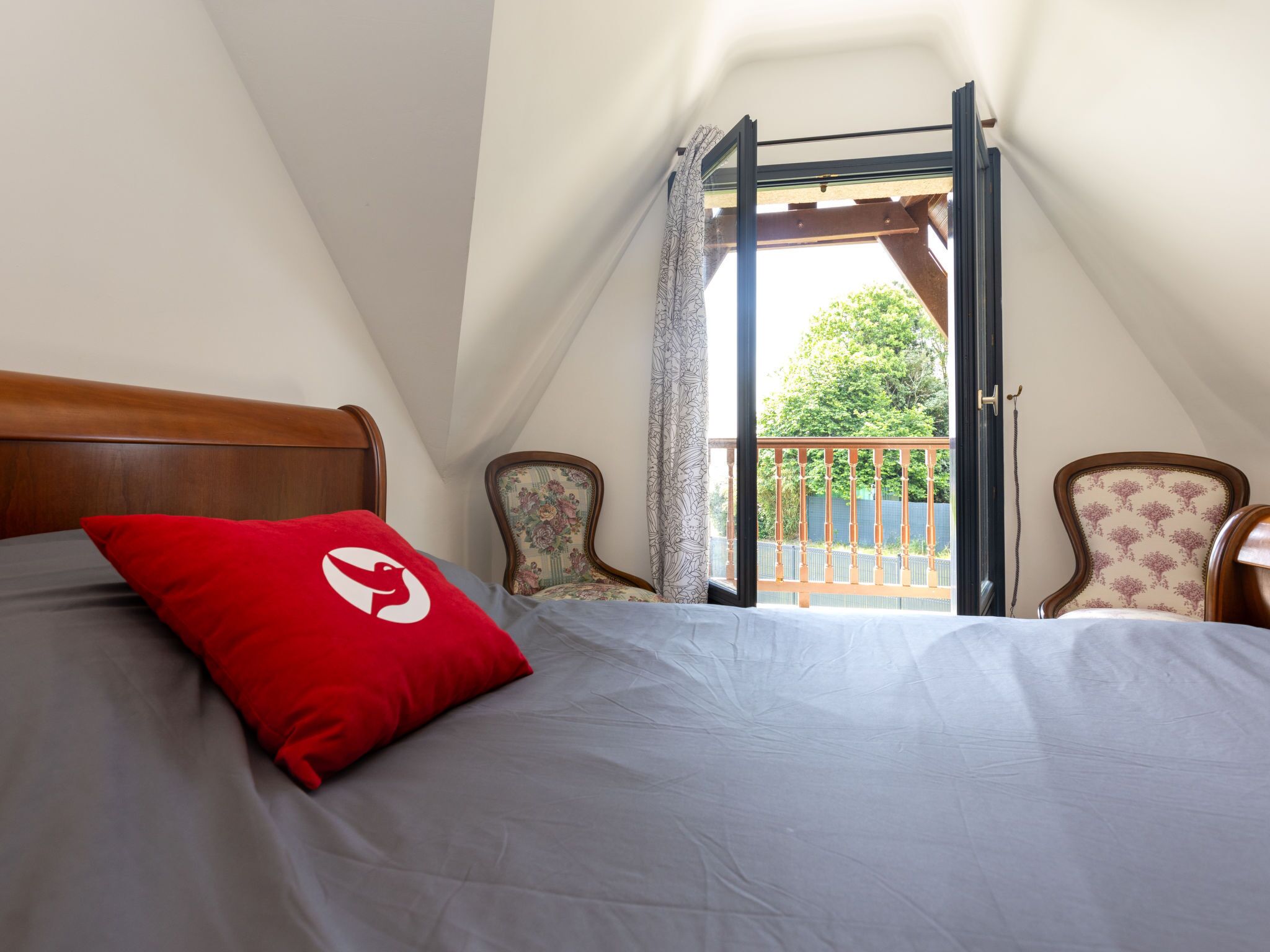 A bedroom with a bed, a red pillow, and a view of a balcony with chairs.