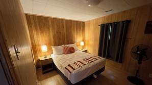 A wooden-paneled bedroom with a bed, two bedside lamps, a standing fan, and a window with curtains. - Charming 2-bedroom house in Devil's Elbow Osgood, IN (Osgood)