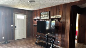 A room with wooden paneling, a flat-screen TV on a stand, a door, and a sign that reads "HOME SWEET HOME". - Charming 2-bedroom house in Devil's Elbow Osgood, IN (Osgood)