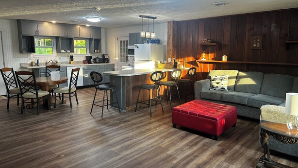 A modern kitchen with a central island, dining area with chairs, and a cozy living room with a sofa and ottoman. - Charming 2-bedroom house in Devil's Elbow Osgood, IN (Osgood)