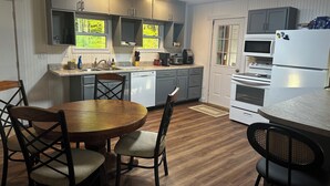 A compact kitchen with a dining area, featuring a round wooden table, chairs, a built-in dishwasher, and a refrigerator. - Charming 2-bedroom house in Devil's Elbow Osgood, IN (Osgood)