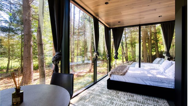 A modern bedroom with a large bed, a dining table, and a view of a forest through large windows. - MIRROR CABIN IN THE FOREST WITH HOT TUB (Windham)