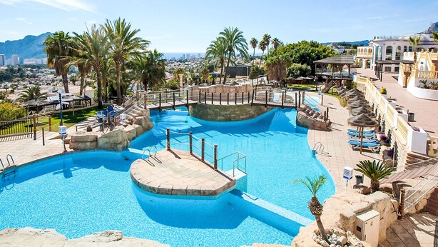 Una gran piscina al aire libre con varias secciones, rodeada de palmeras y un paisaje urbano de fondo.