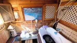 A wooden cabin interior with a bathtub, wooden benches, a window with a view, and a lattice wall.