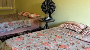 A room with two beds, each with floral-patterned bedding. There is a small table with a fan and a power strip.