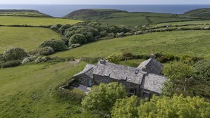 A stone house with a sloped roof, surrounded by rolling green hills and a view of the sea.