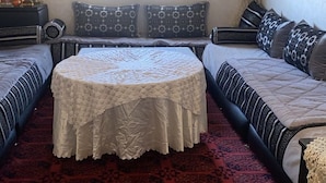 Een kamer met een bed, een ronde tafel met een kantkleed, een bank met gestreepte kussens, en een rood tapijt met dessin.
