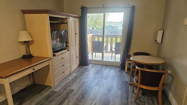 Una habitación con escritorio de madera, televisión, mesa de comedor con sillas y puerta corrediza de vidrio que da a un espacio exterior con sillas.