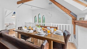 Uma área de refeições com uma mesa de madeira posta para o café da manhã, paredes brancas e teto inclinado com vigas aparentes.
