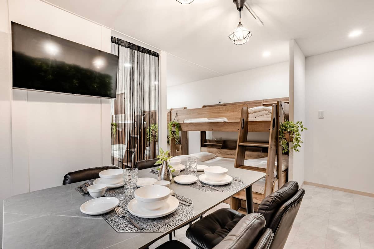 A modern dining area with a large table set for four, a TV mounted on the wall, and a bunk bed structure in the background.