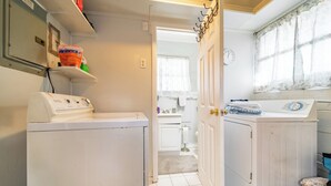 A laundry room with two washing machines, a shelf with cleaning supplies, and a door leading to a bathroom.