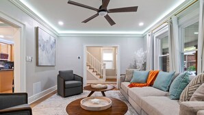 A modern living room with a ceiling fan, a sofa, a coffee table, and a dining area visible through an open door. - Spacious Pittsburgh Home with Deck and Grill (Pittsburgh)