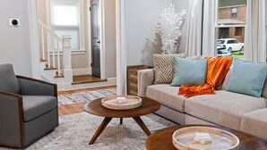 A living room with a grey armchair, a beige sofa with orange and blue cushions, and a wooden coffee table with a tray. - Spacious Pittsburgh Home with Deck and Grill (Pittsburgh)