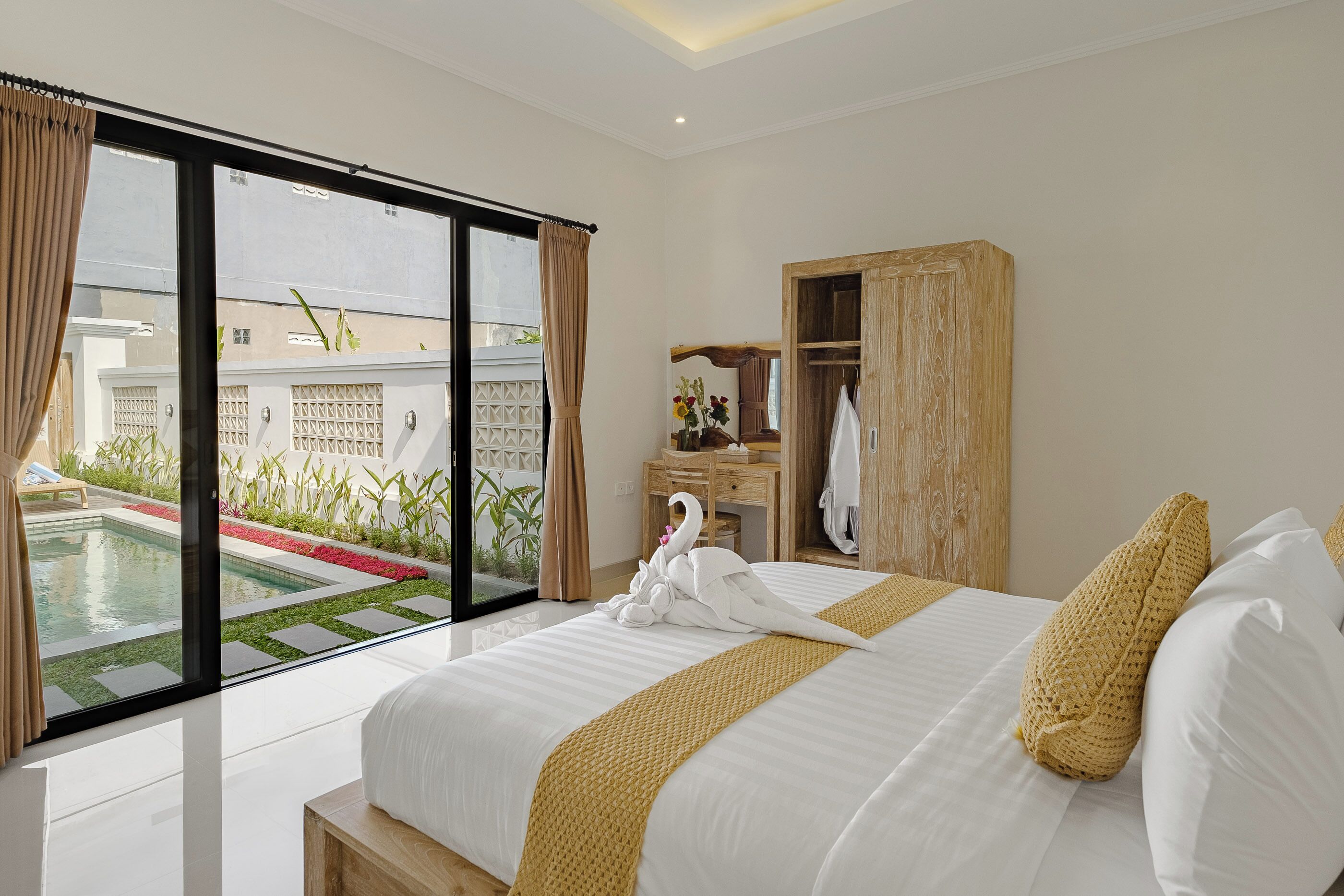 Quarto de hotel moderno com uma cama grande, um guarda-roupa de madeira, uma mesa de escritório e vista para uma piscina e jardim.
