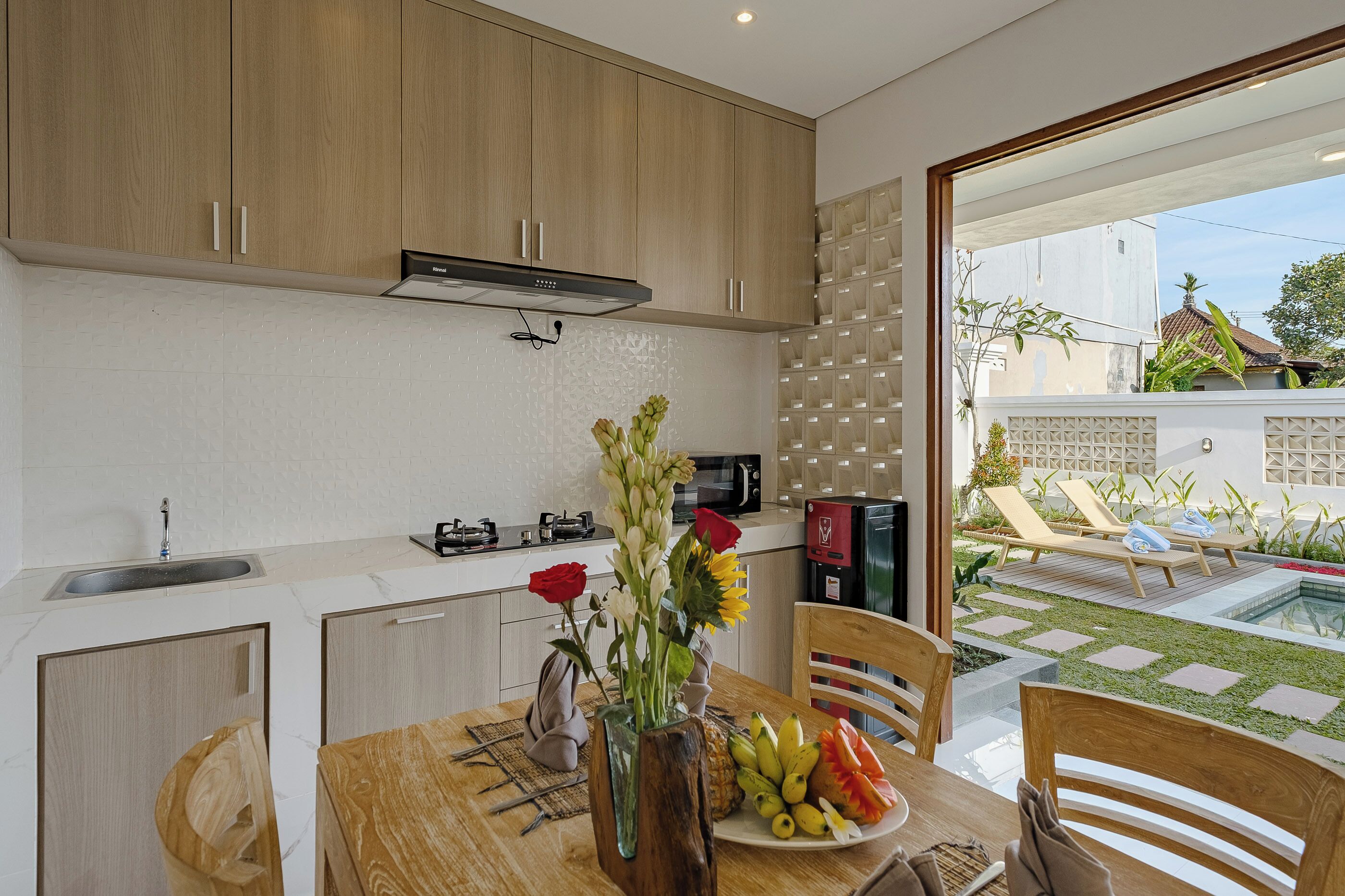 A modern kitchen with wooden cabinets, a dining table with a vase of flowers, and a view of a backyard with a pool and lounge chairs.