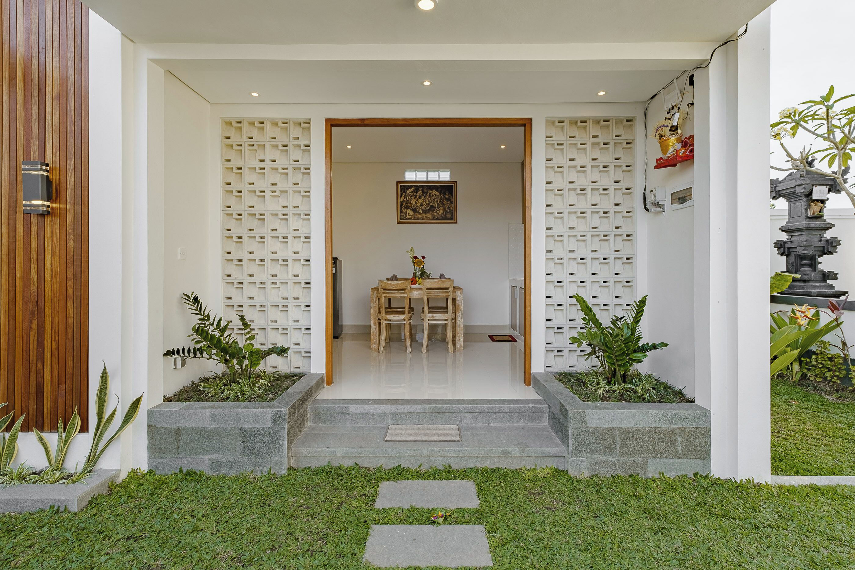Une entrée moderne avec une porte en bois, un chemin de pierre menant à un espace détente et un mur blanc orné d’un motif en quadrillage.