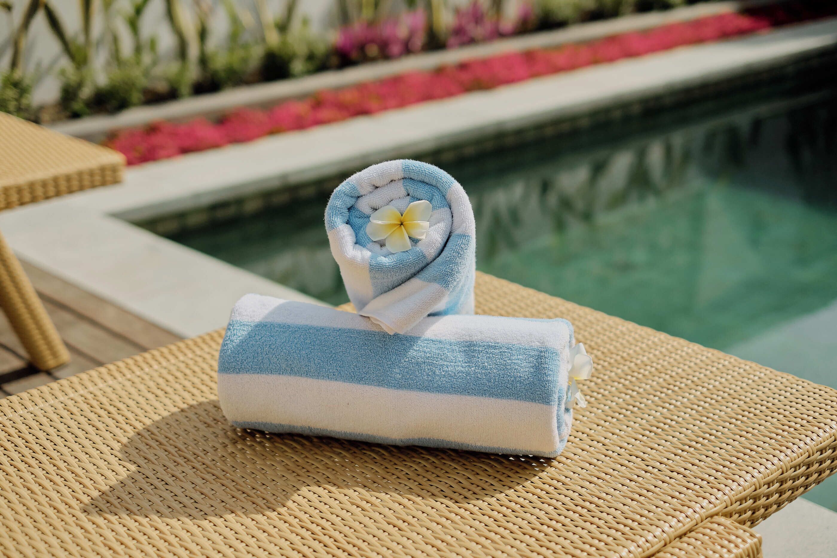 Une serviette bleue et blanche, ornée d’une fleur, est posée sur une chaise en osier près d’une piscine.