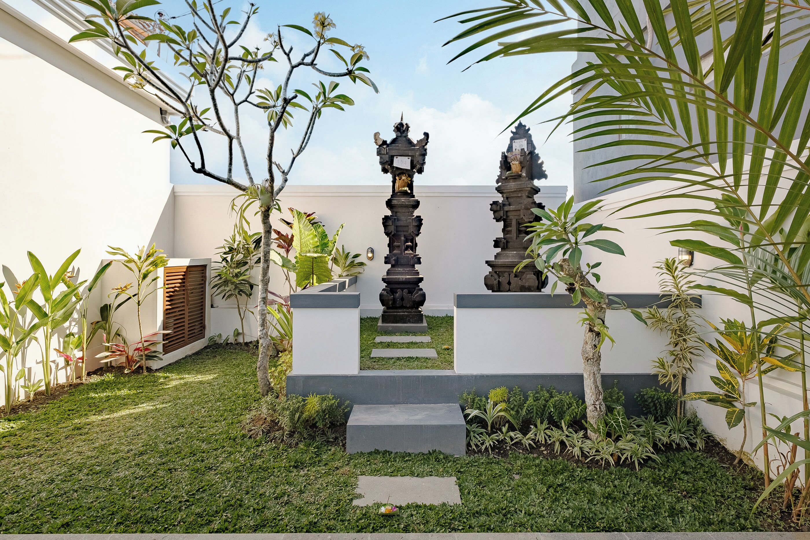 A courtyard with a central stone structure, surrounded by various plants and a white building.
