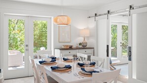 Un comedor luminoso con una mesa blanca preparada para cuatro personas, rodeada de sillas blancas. Hay una lámpara colgante sobre la mesa y un cuadro en la pared.