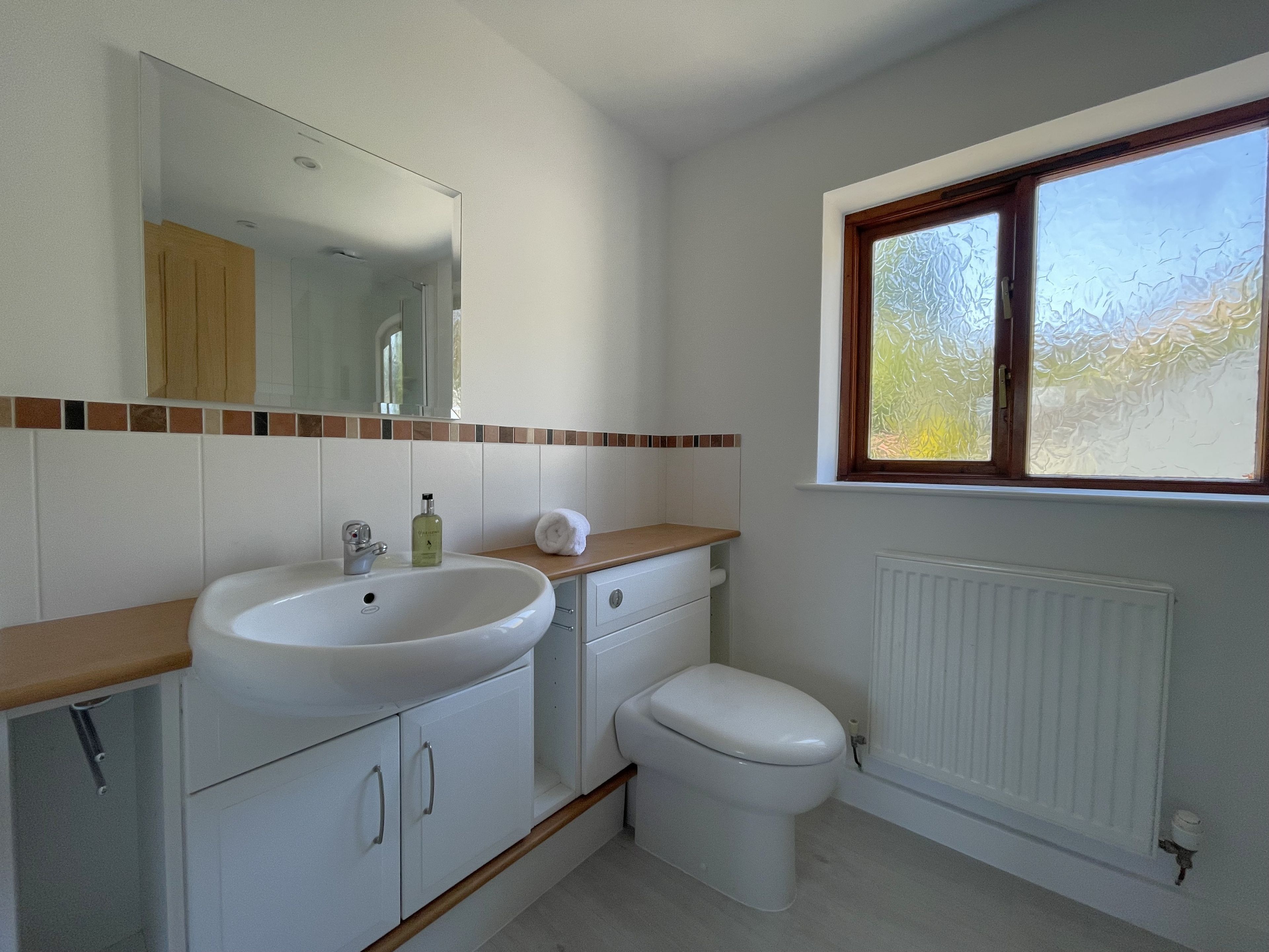 Une salle de bain avec un lavabo blanc, un miroir et des toilettes. Il y a une fenêtre avec du verre dépoli et une vue sur la verdure à l’extérieur.
