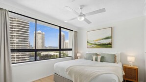 A modern hotel room with a large window offering a city view, a bed with a white comforter, a ceiling fan, and a bedside table with a lamp. - Carrington Family Beach Getaway - Ocean View, WiFi (Main Beach)
