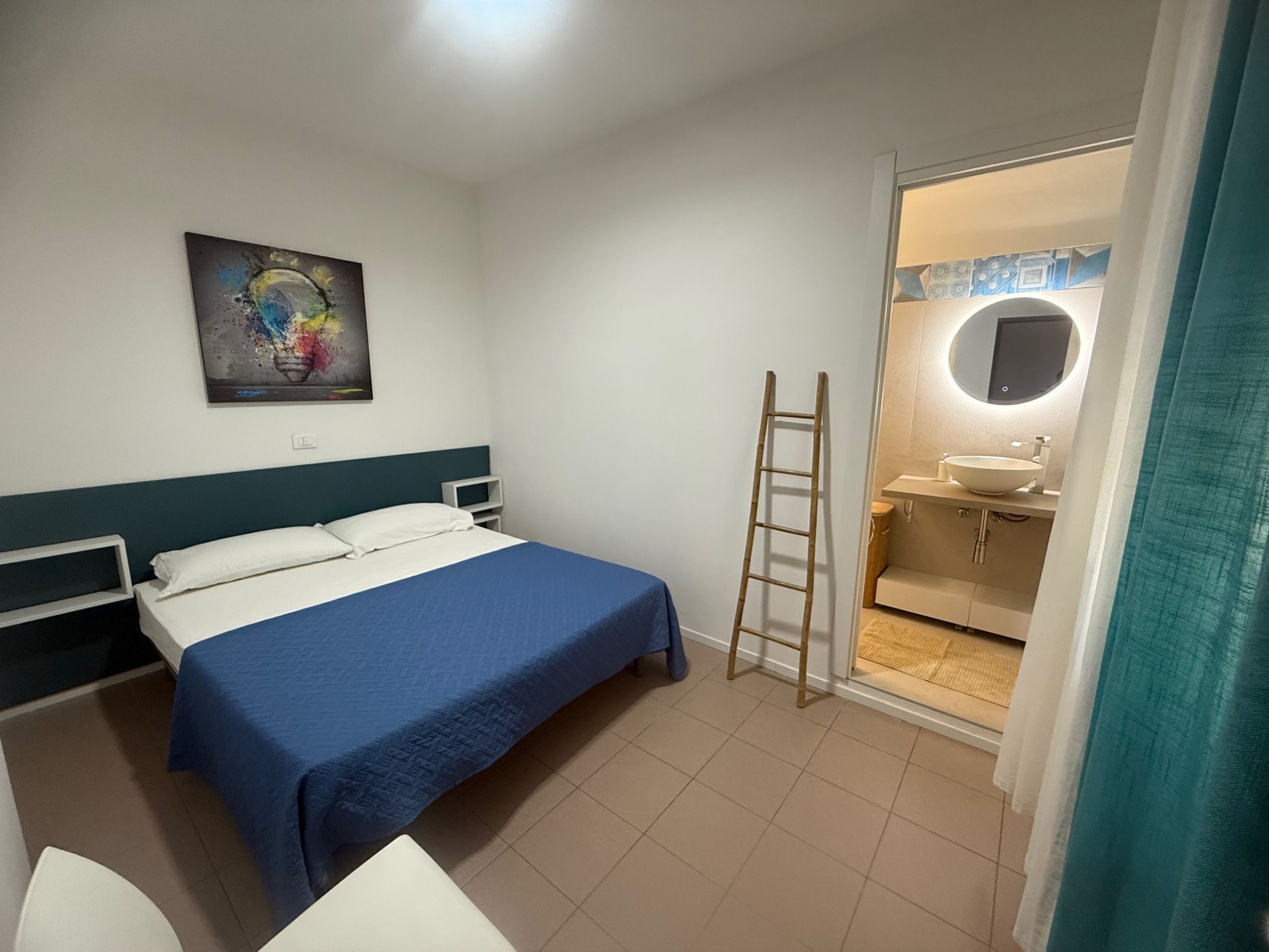 Une chambre d’hôtel avec un lit, une table de chevet, un miroir et une salle de bain visible à travers une porte ouverte.