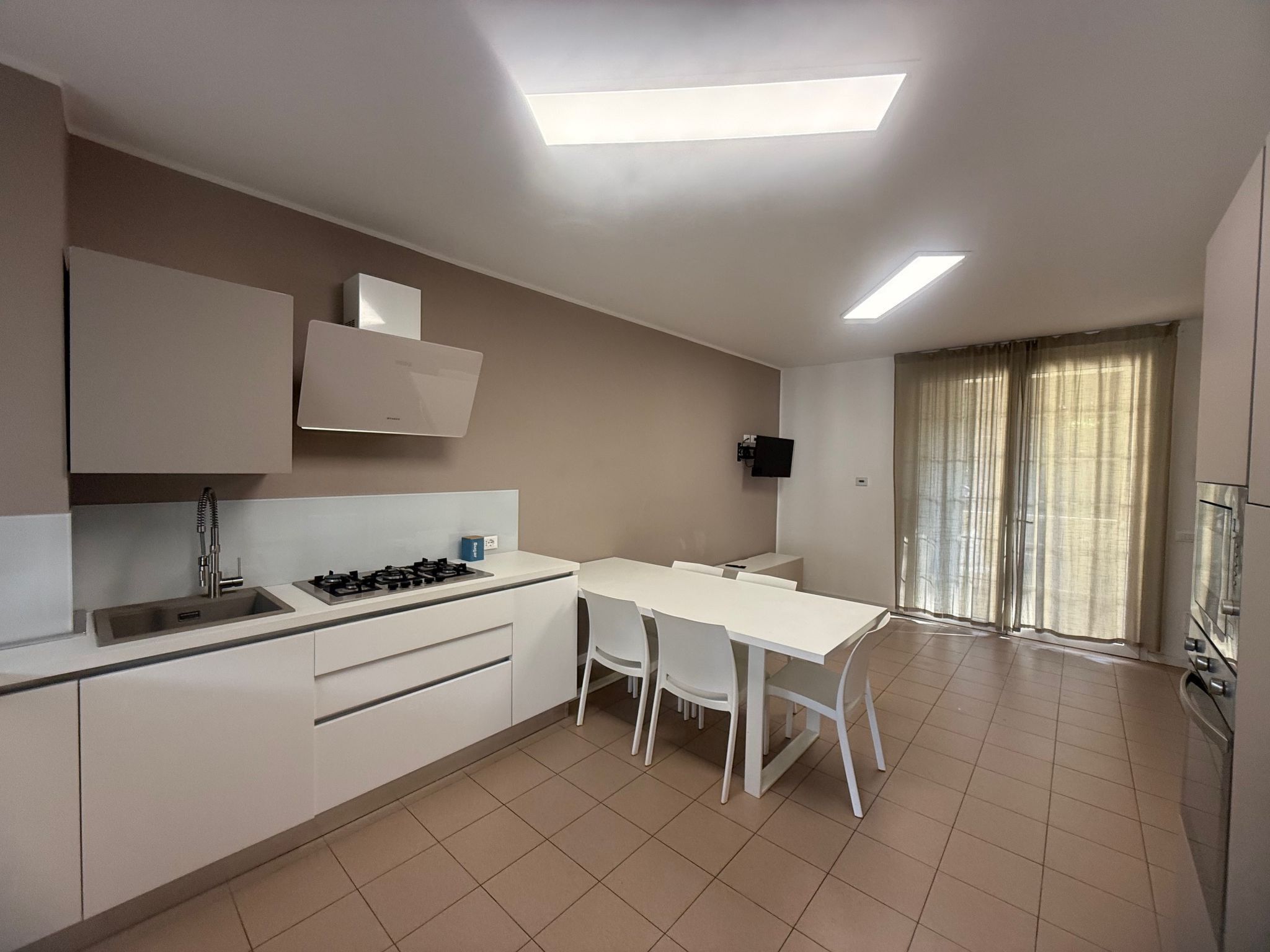 Una cucina moderna con piano cottura a gas, lavello e pensili bianchi. Si nota un tavolo da pranzo con sedie e un televisore fissato alla parete.