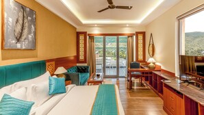 Une chambre d’hôtel avec un lit, un canapé, un bureau et une télévision. Elle dispose d’un balcon avec vue sur les collines.