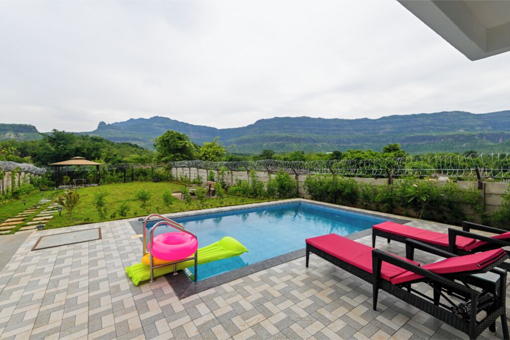 A swimming pool with a view of mountains, surrounded by a garden and a patio area with lounge chairs.