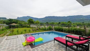 A swimming pool with a view of mountains, surrounded by a garden and a patio area with lounge chairs.