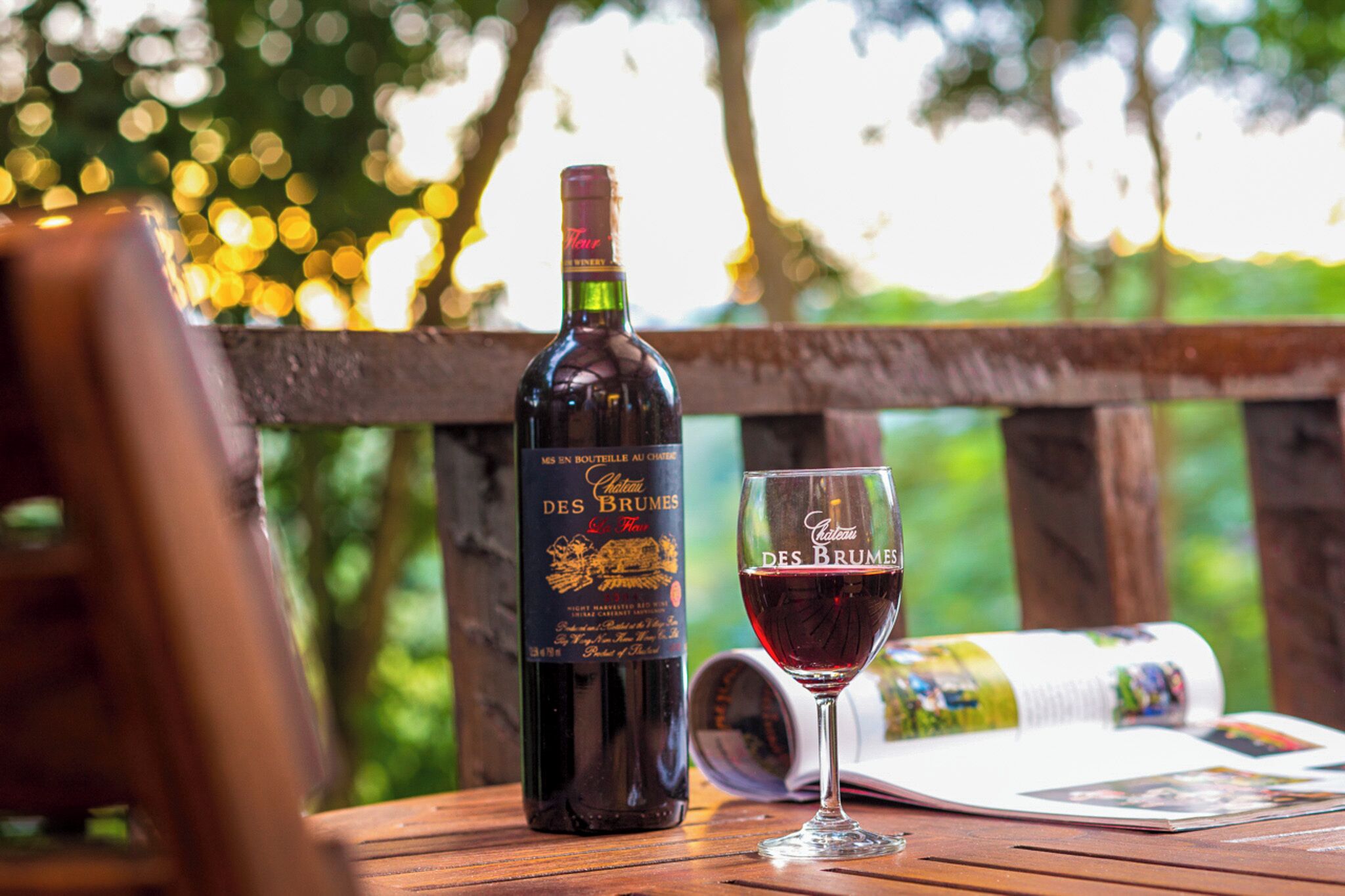 A bottle of wine and a glass on a wooden table with a magazine, set outdoors with a blurred natural background.