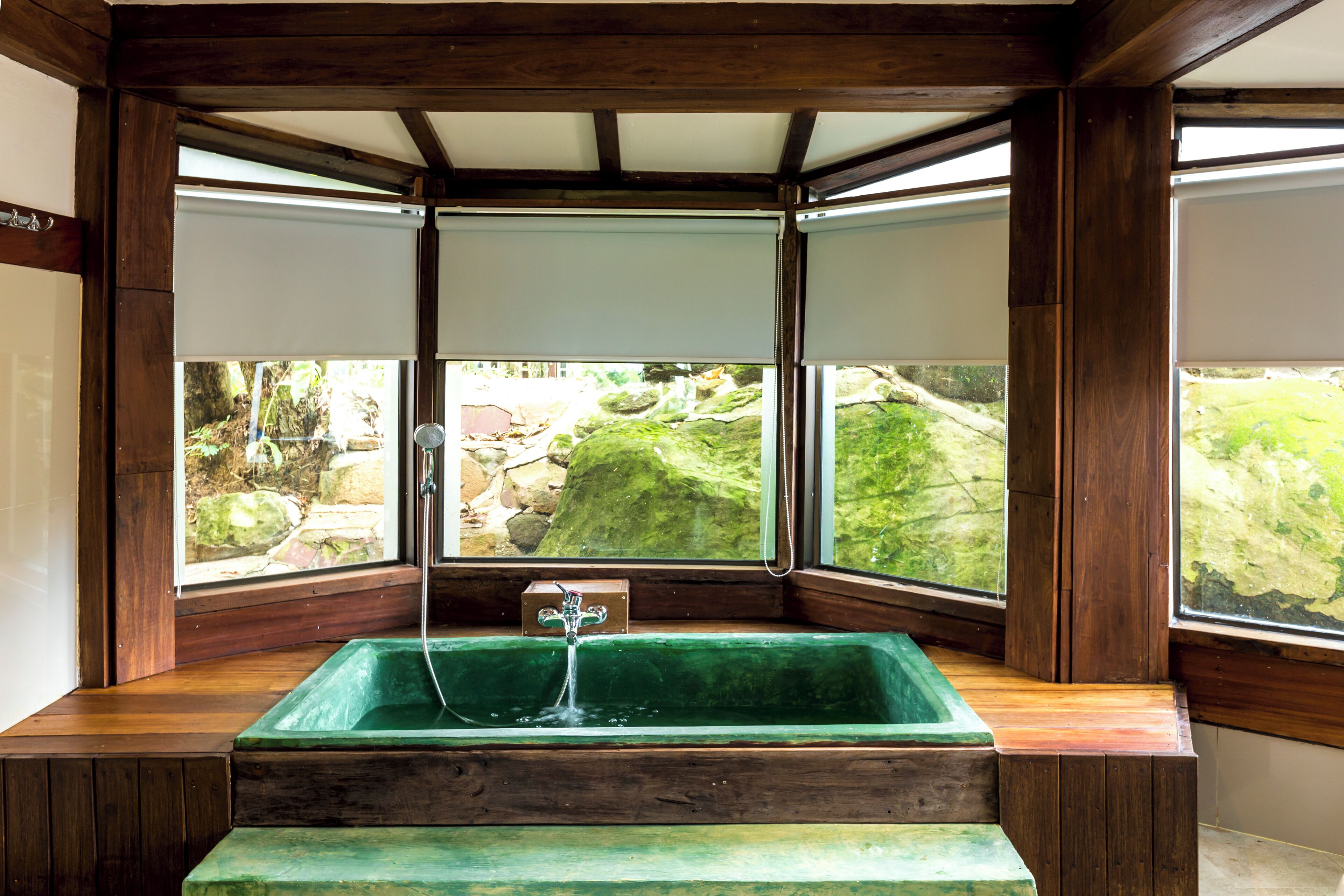 A room with a green bathtub, wooden beams, and large windows overlooking a garden.