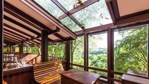 Una terraza de madera con un banco y una mesa, amplias ventanas con vistas al bosque.